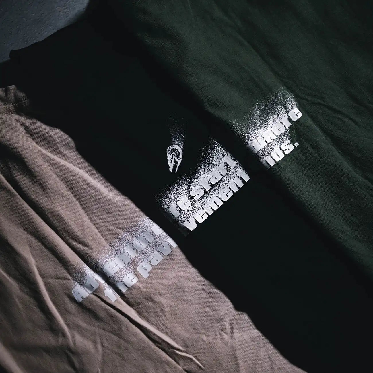 Close-up lifestyle shot of Pavement Ends T-Shirts in Tan, Black, and Olive laid out flat, showcasing spray-fade graphic design.