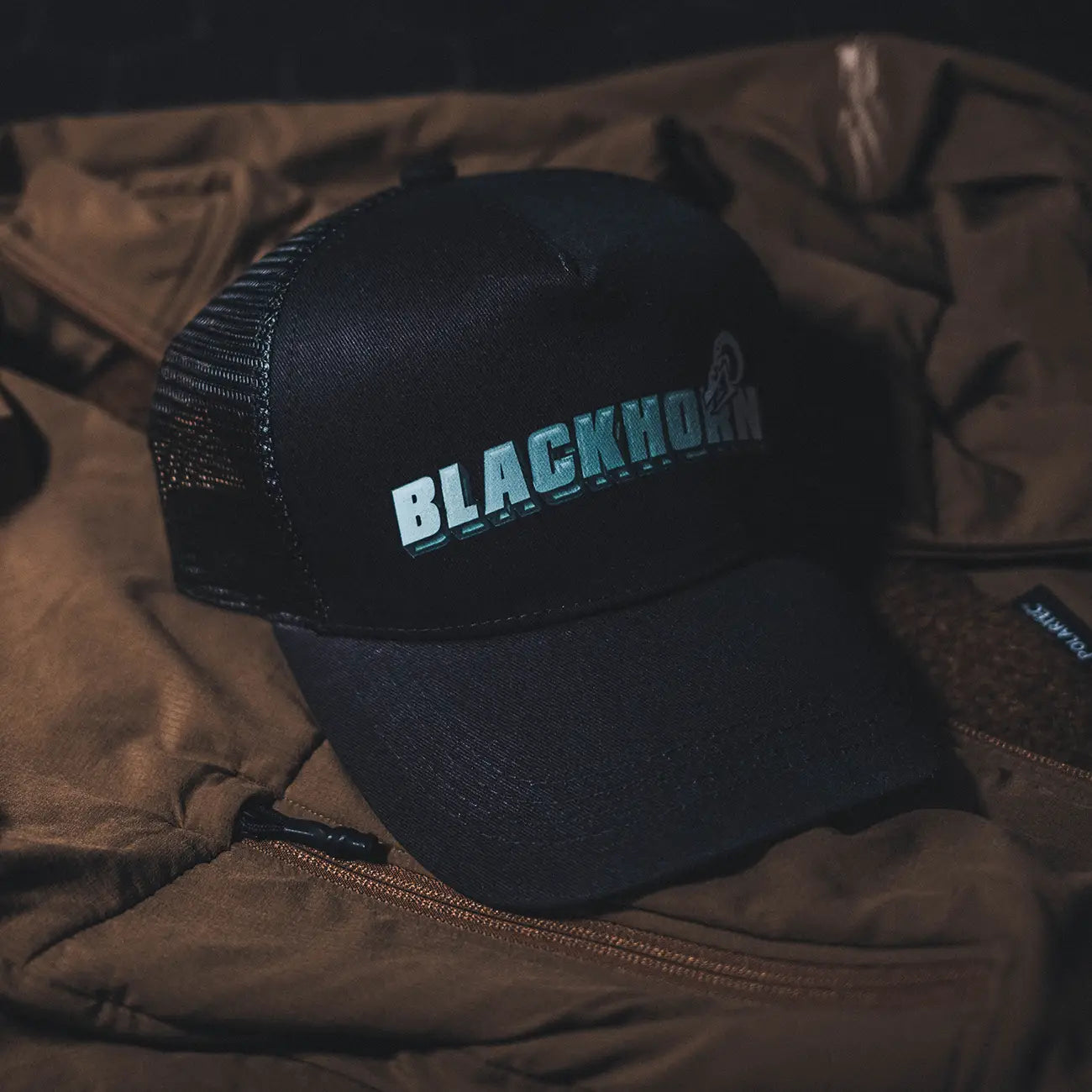 Blackhorn Polymark Trucker Hat in black with acrylic effect logo, displayed on a rugged brown field jacket in low-light garage setting.